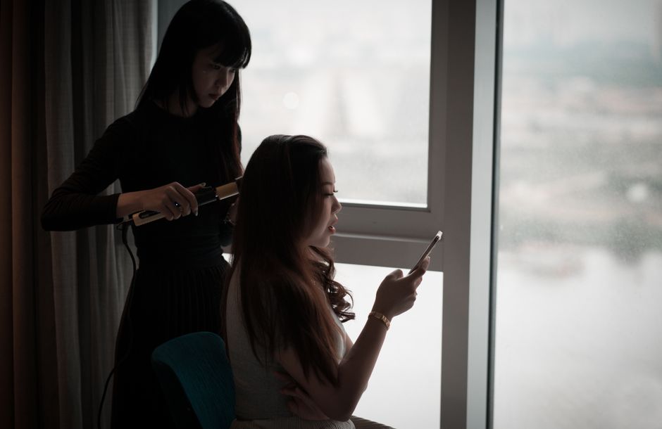 Asian woman curls hair while using a smartphone by a window in dim light.