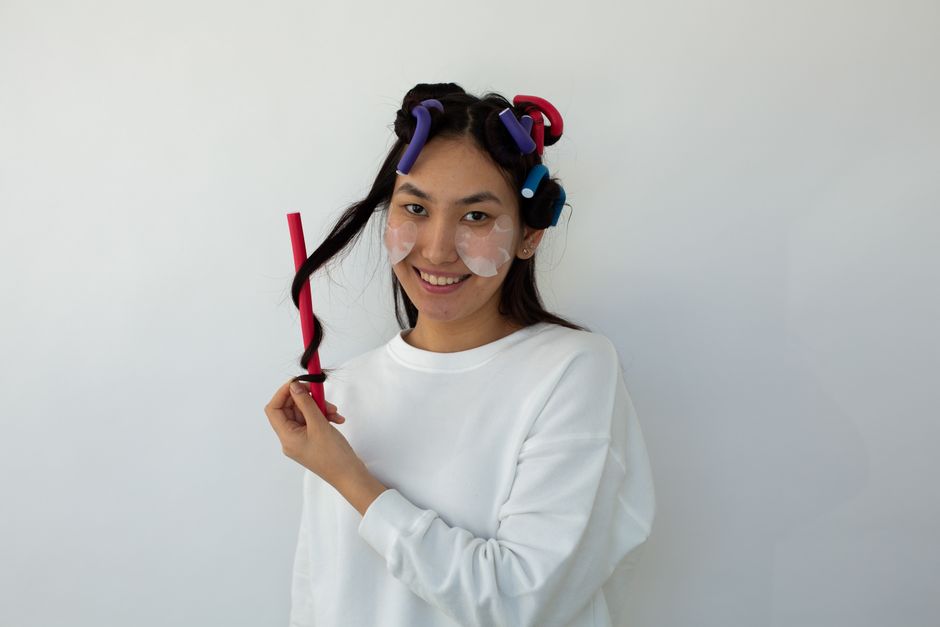 Content Asian female with moisturizing eye patches on cheeks and hair rollers looking at camera on white background