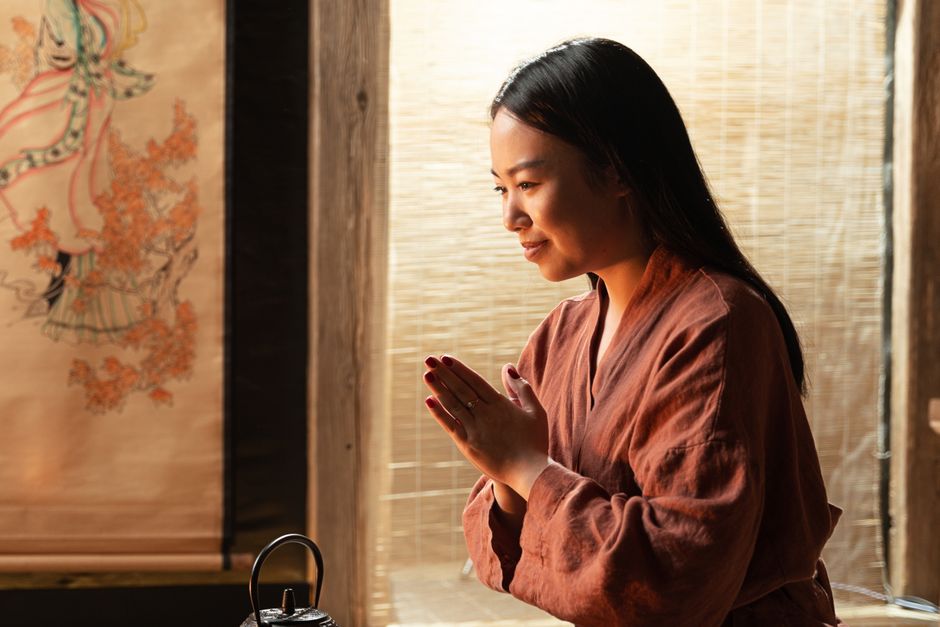 An Asian woman in a traditional outfit with hands clasped in prayer indoors.